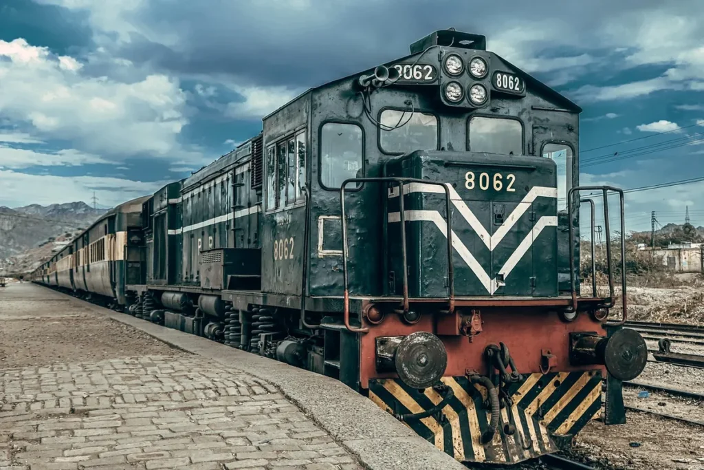 A green Pakistan Railways diesel locomotive standing beside a brick station platform under a cloudy sky.