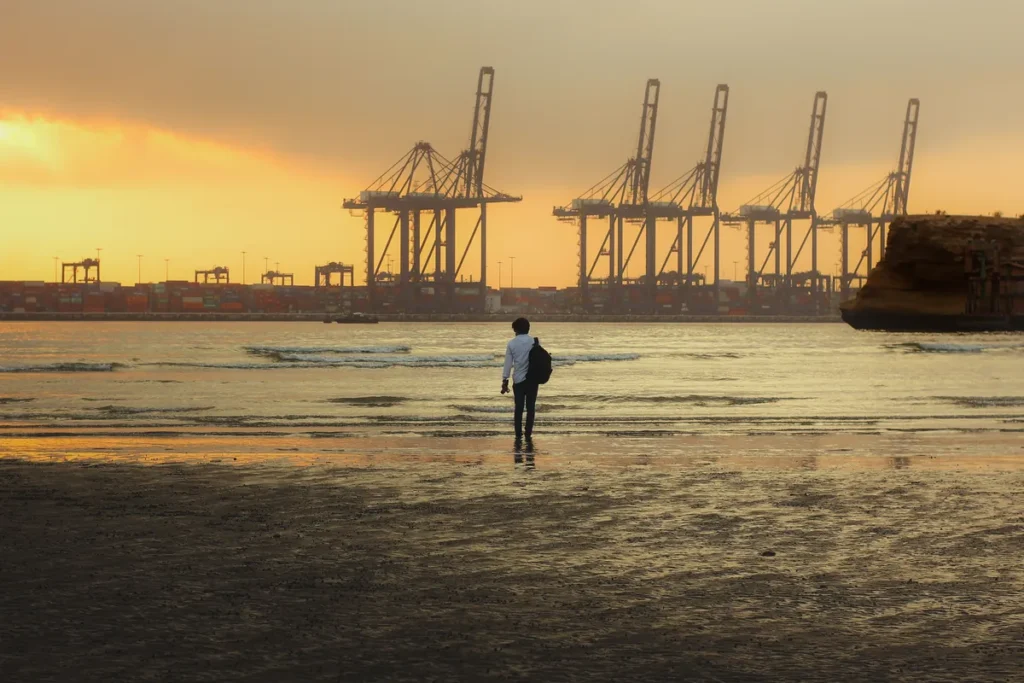 A man walking on Karachi beach at sunset, with the port's container cranes silhouetted behind him.