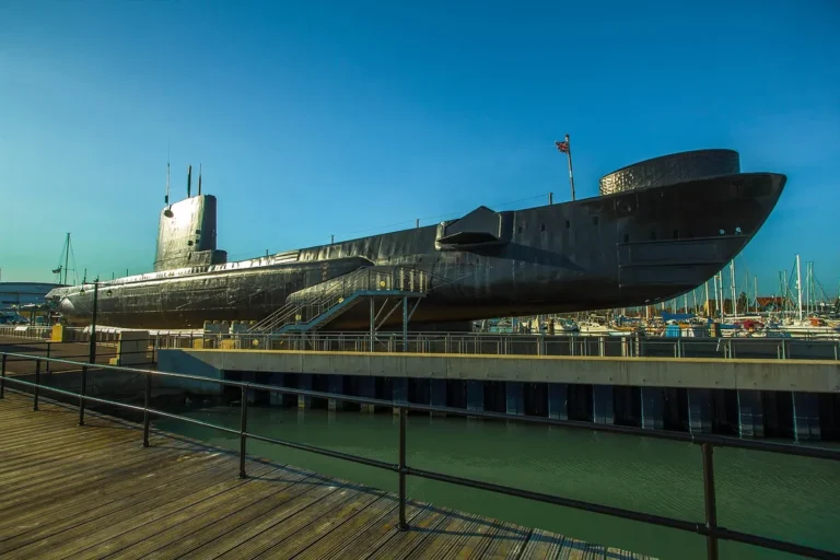 A black submarine displayed on land beside a harbour boardwalk as a museum exhibit, under a blue sky.