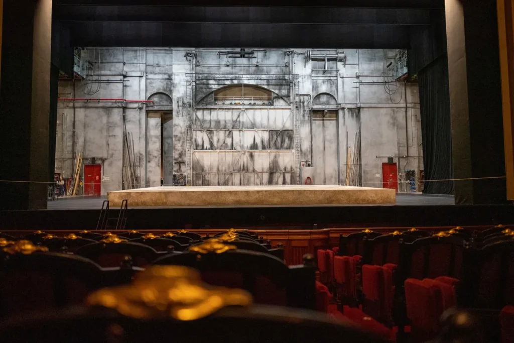 An empty theatre stage with a painted backdrop, seen across rows of red auditorium seats.