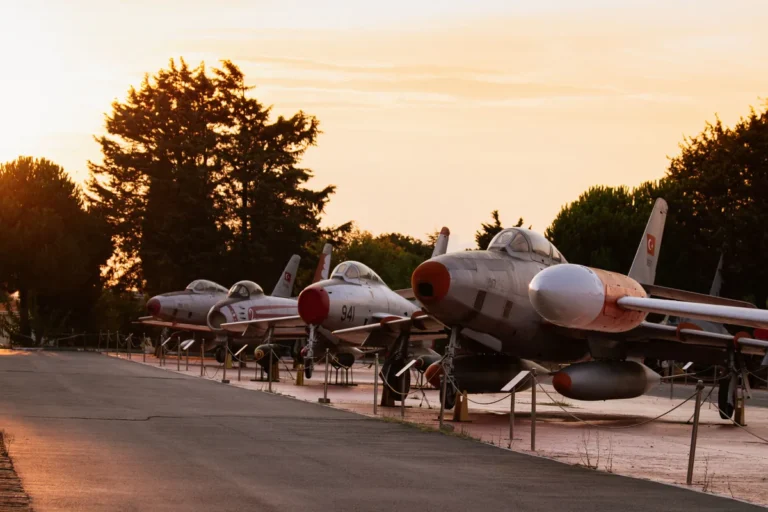 A row of vintage military jet fighters on outdoor display beside a paved road at sunset.
