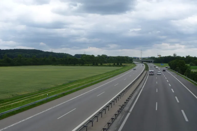 A divided motorway with light traffic running between green fields and woodland under a cloudy sky.